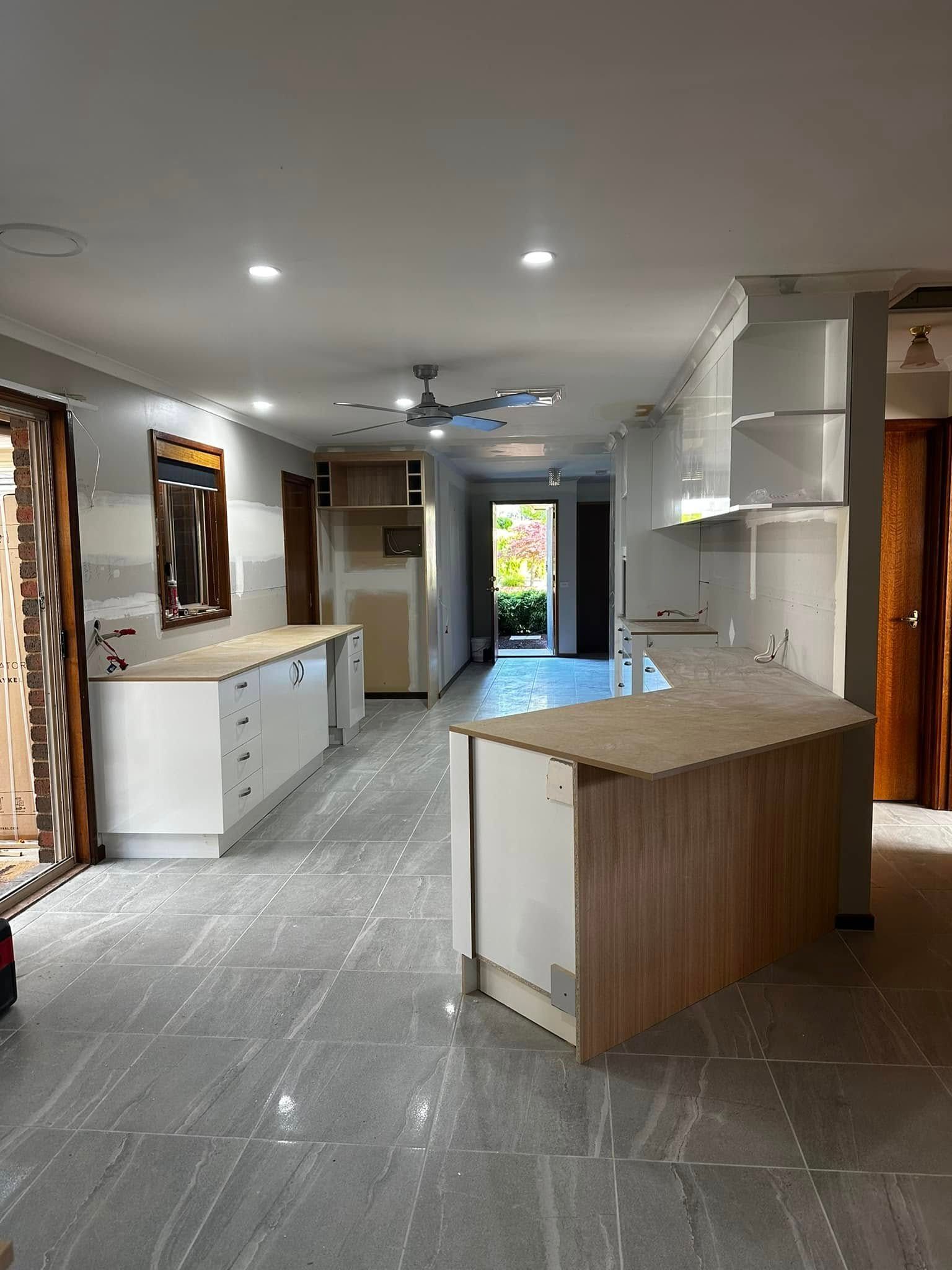 An open-plan kitchen with white cabinetry and light wood countertops. The space is under construction, with tile floors and a view to the exterior.