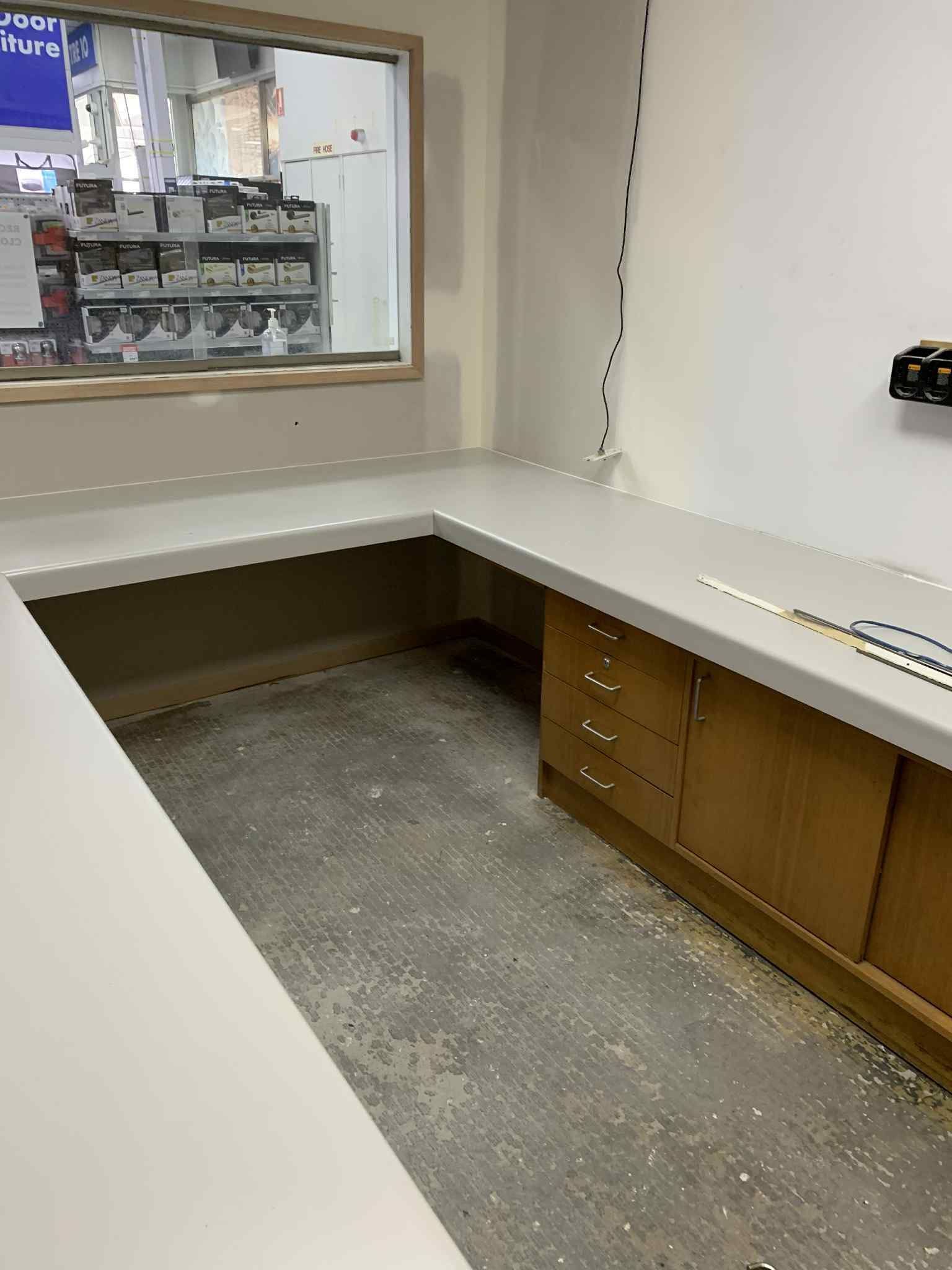 L-shaped counter in a room with a window overlooking a store. Cabinets sit below the counter. The floor is unfinished concrete.
