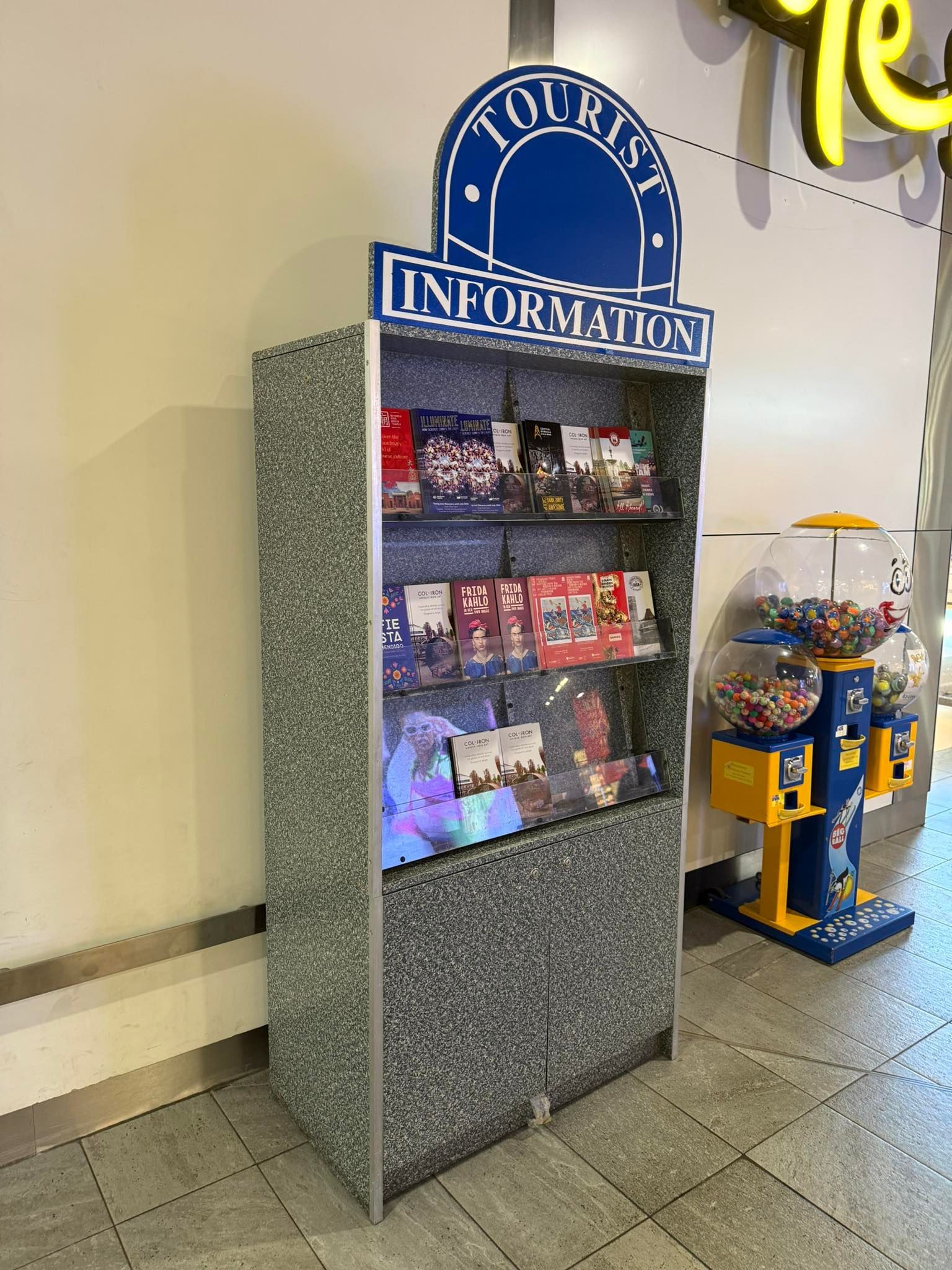 Tourist Information display case with brochures, next to a gumball machine, inside a brightly lit area.