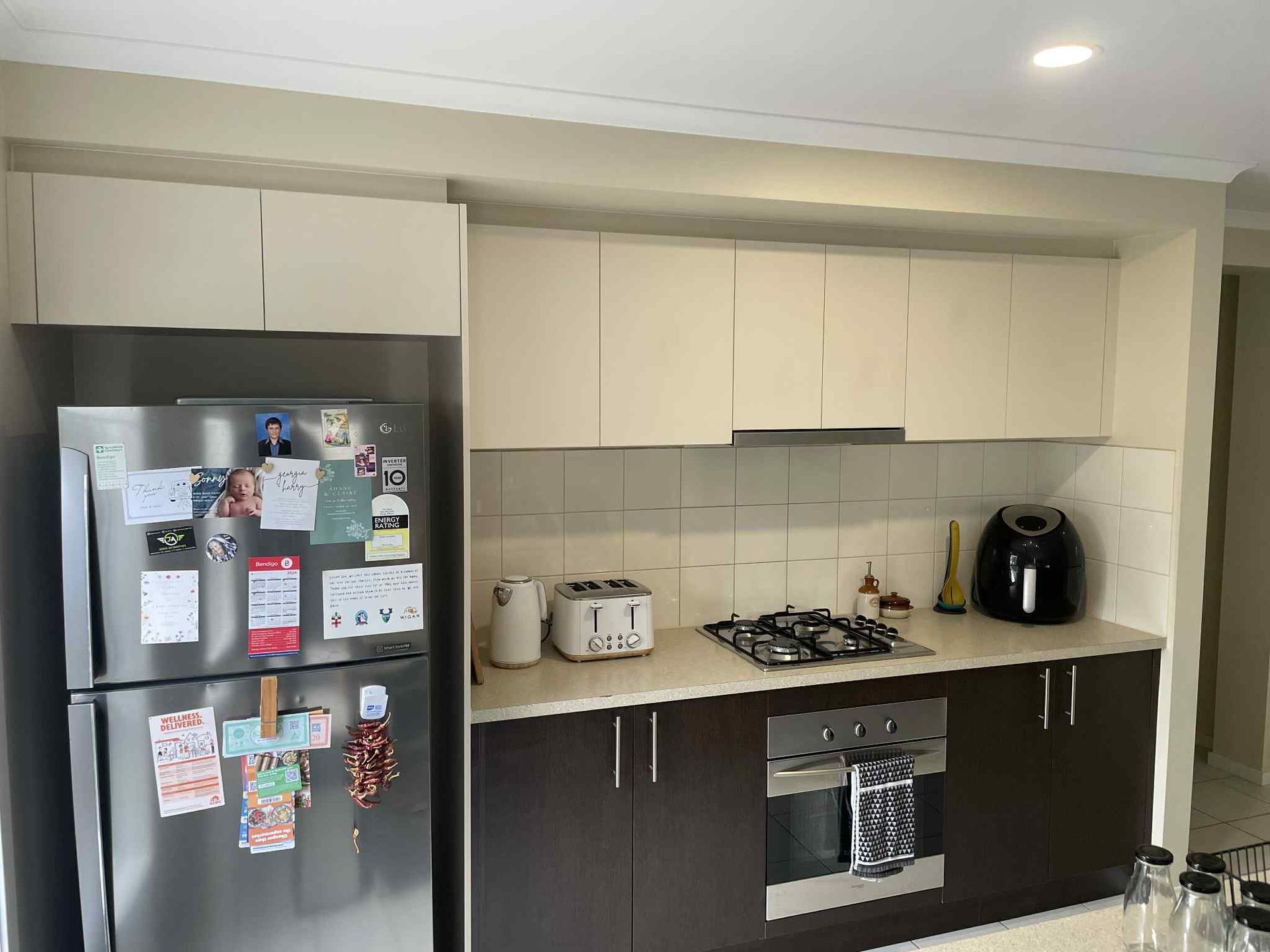 A modern kitchen with a stainless steel fridge, cream upper cabinets, and brown lower cabinets. Appliances include a gas stove and an air fryer.