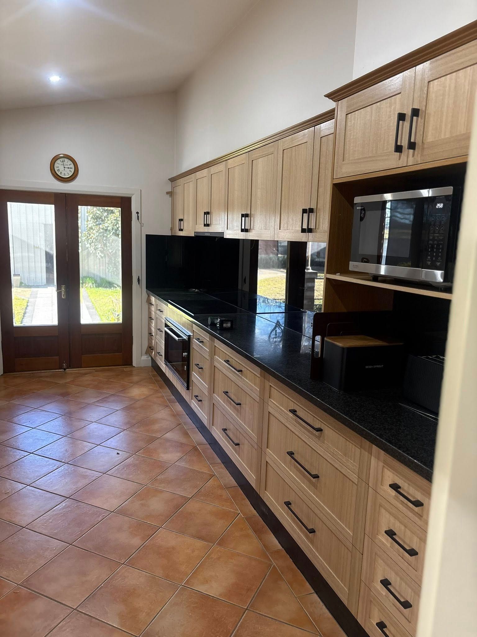 A kitchen with light wood cabinets, black countertops, and a microwave. A doorway leads to the outdoors.
