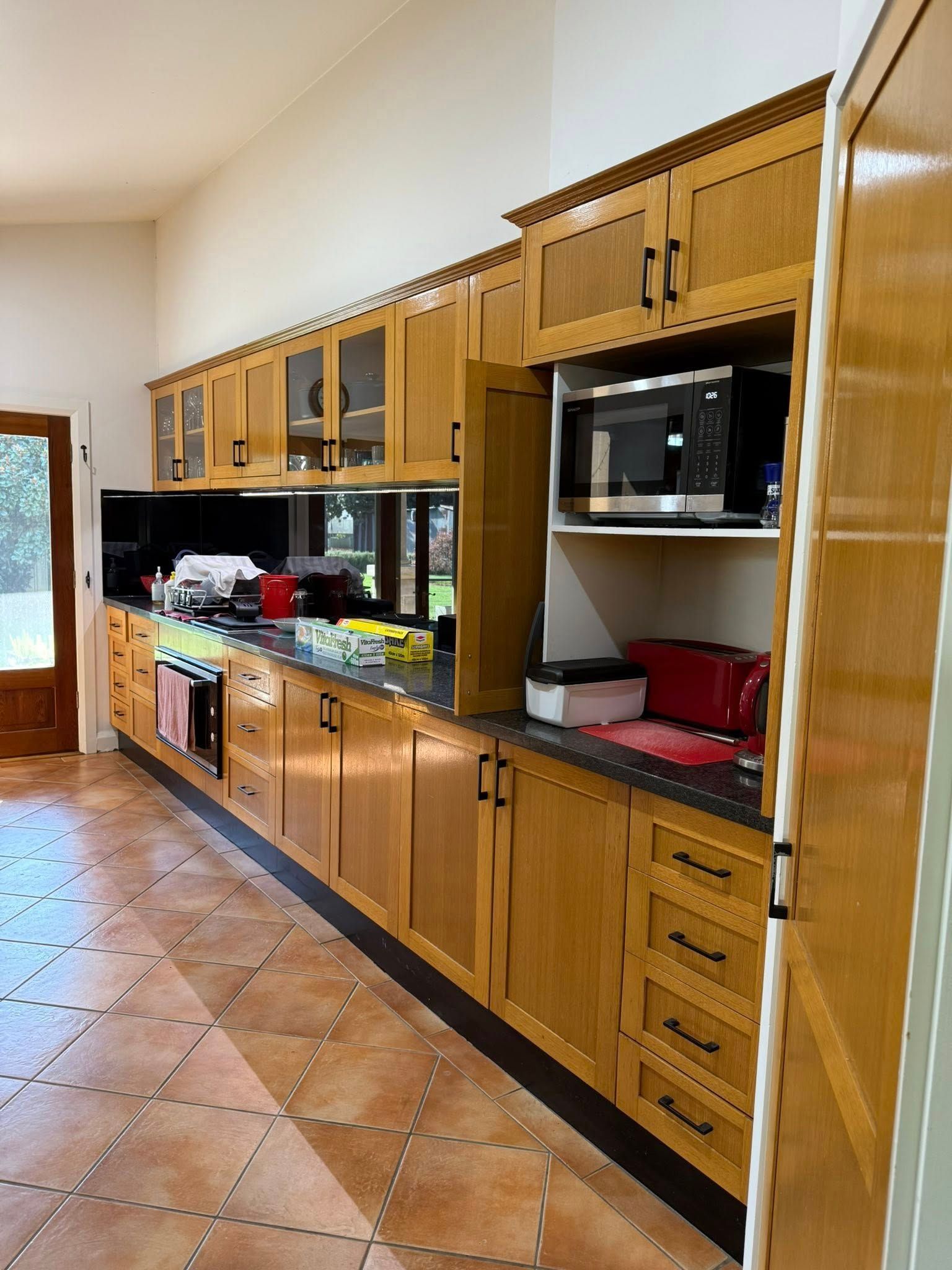 Long, wooden kitchen with upper and lower cabinets. A microwave sits in a cabinet. Dark countertops, brown tiled floor.