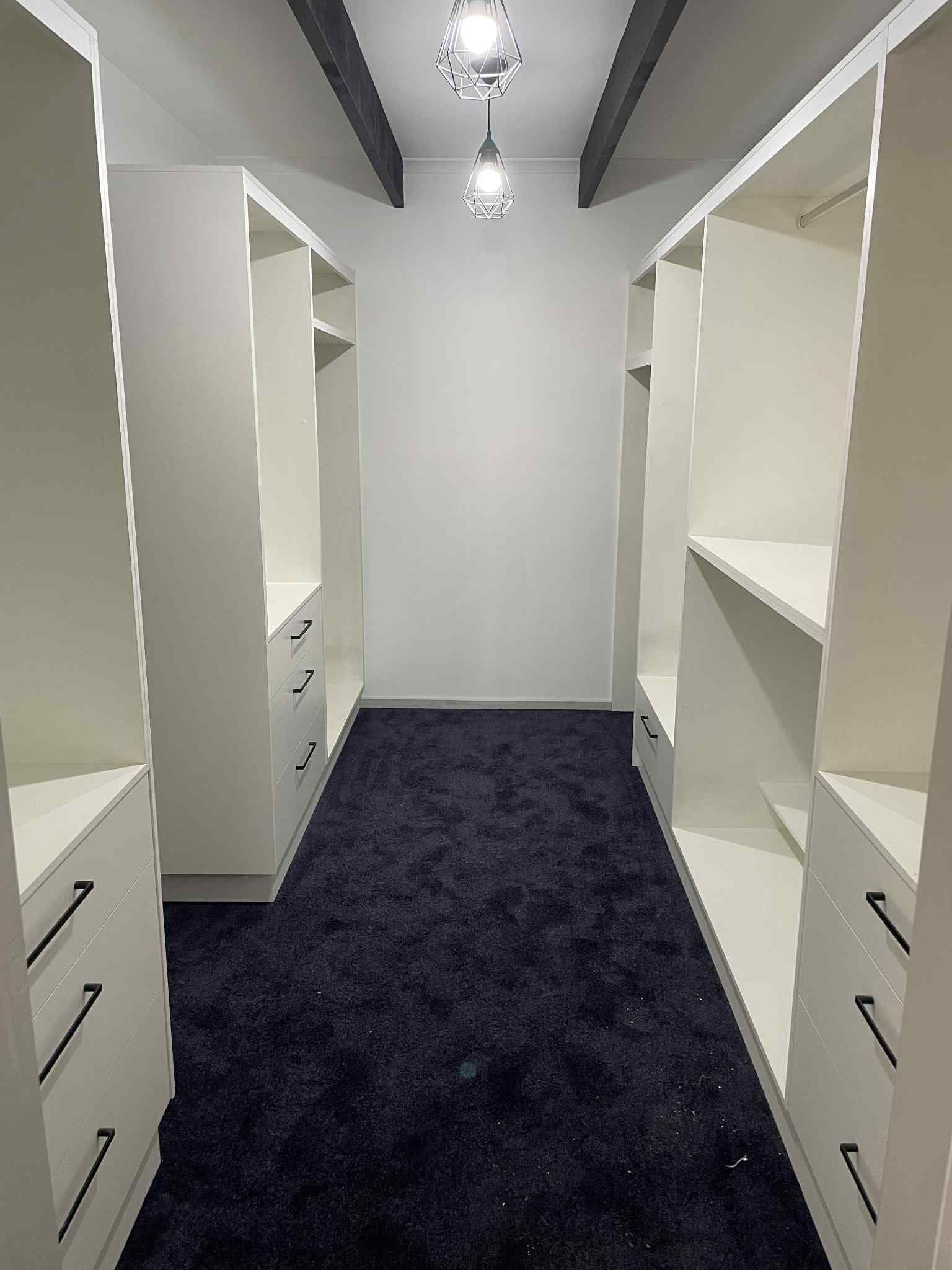 A walk-in closet with white shelving units and black drawer pulls, facing each other with a dark blue carpet. Two lights hang from the ceiling.