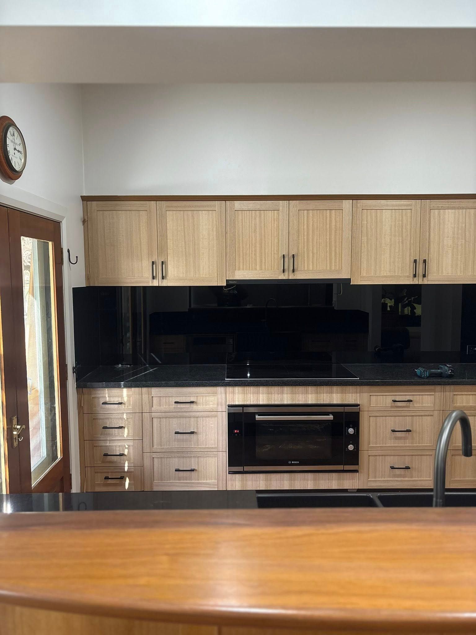 Kitchen with light wood cabinets, black backsplash, oven, and countertop. A clock hangs on the wall, and a glimpse of a door is visible.