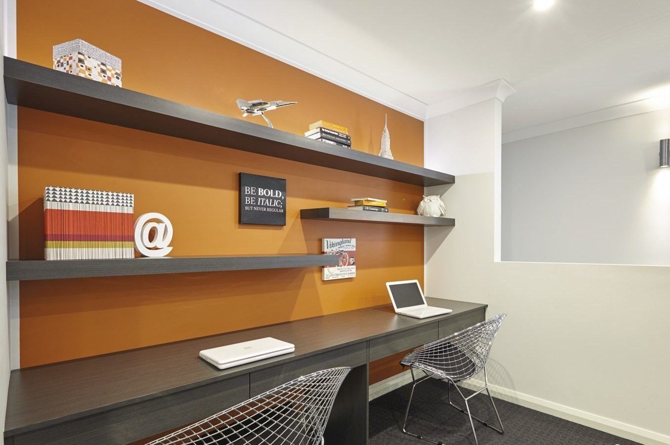 A home office with two built-in desks, floating shelves, and an orange accent wall. Two wire chairs are at the desks, one with a laptop open.