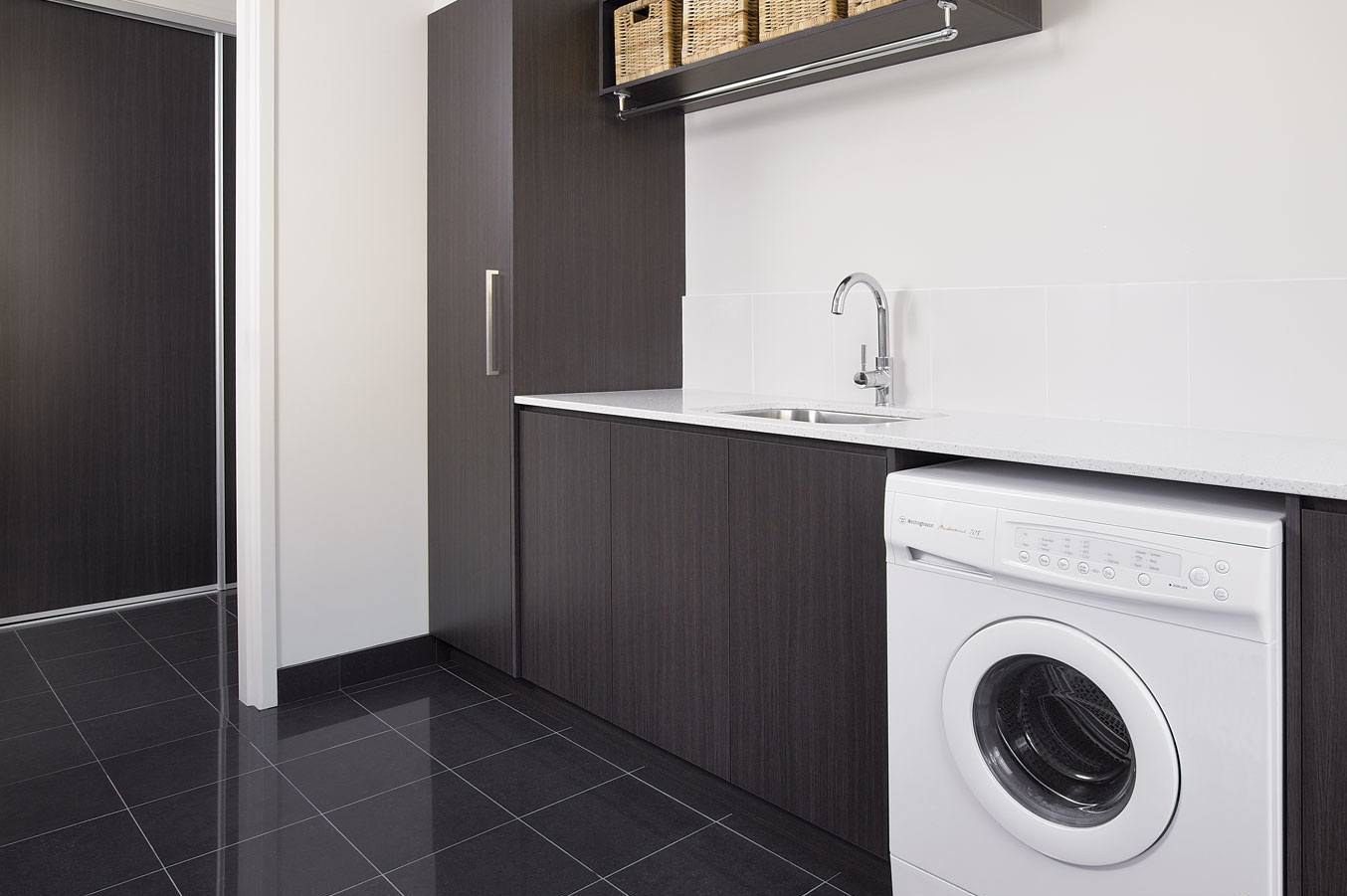 A modern laundry room with dark cabinets, white countertop, and a washing machine. A sink and storage shelves are also present.