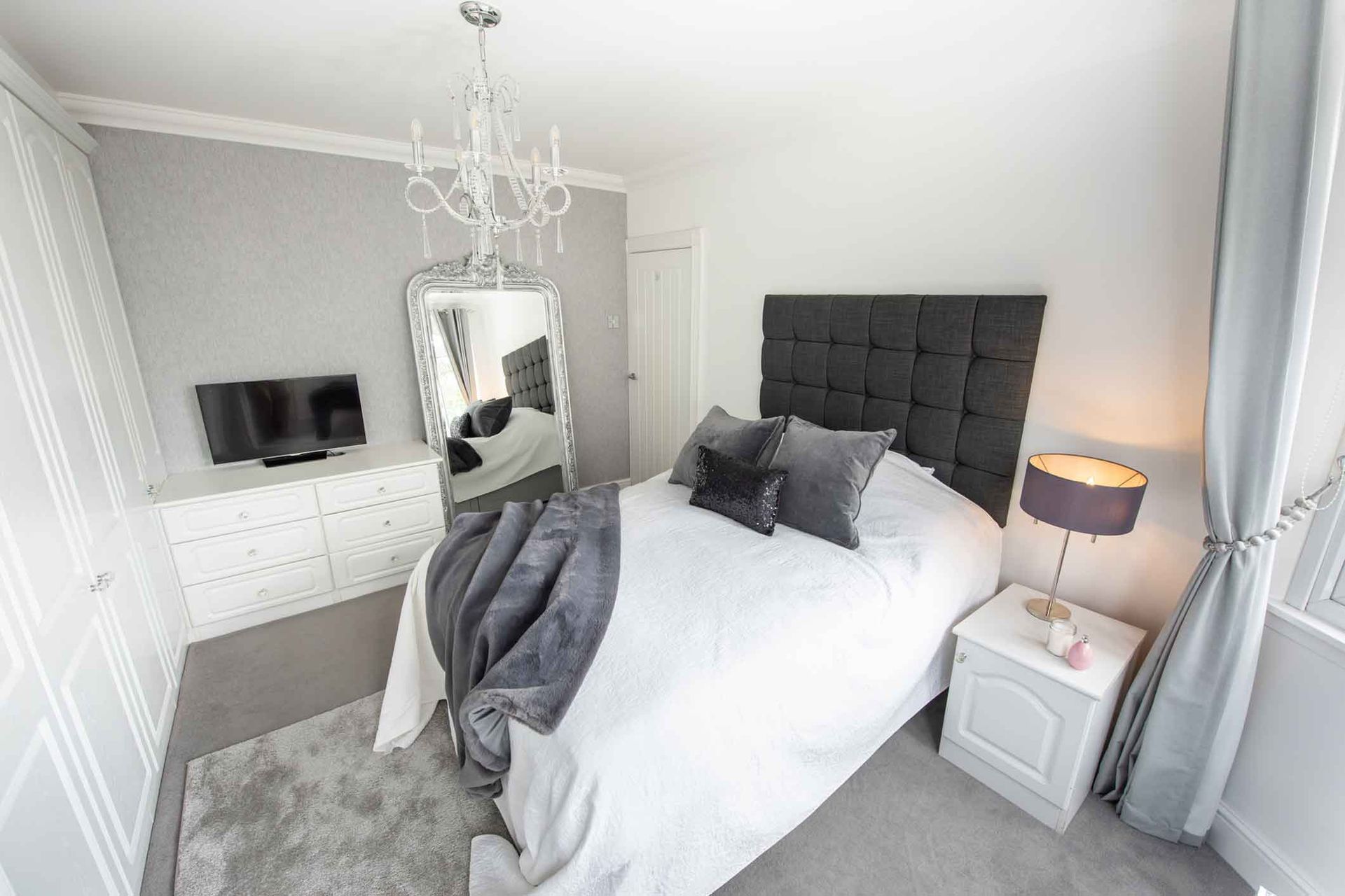 A modern bedroom with a bed, dresser, closet, and mirror. The room has gray and white accents, a chandelier, and a TV.