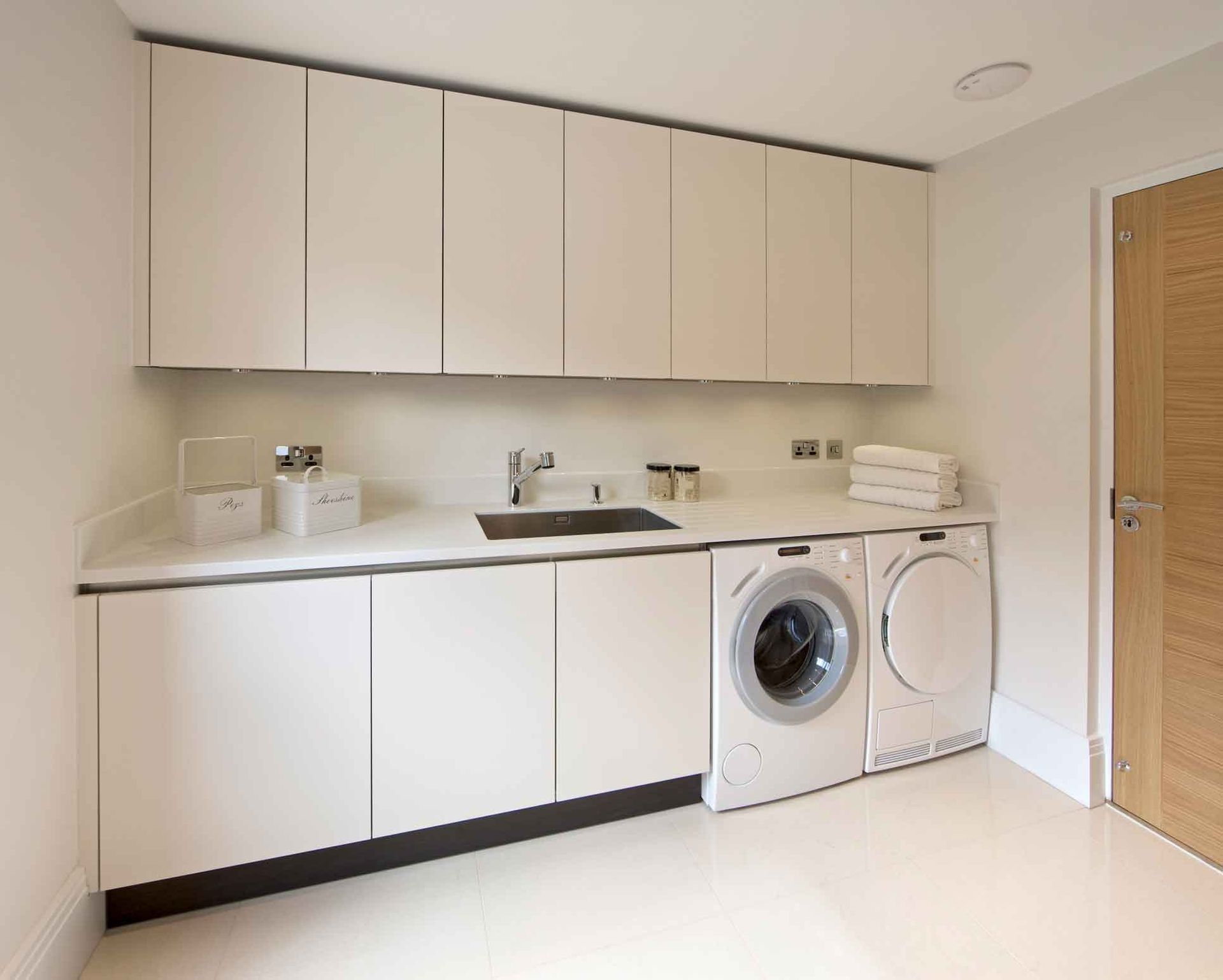 A modern laundry room with white cabinets, a sink, washer, and dryer. Light wood door on the right.
