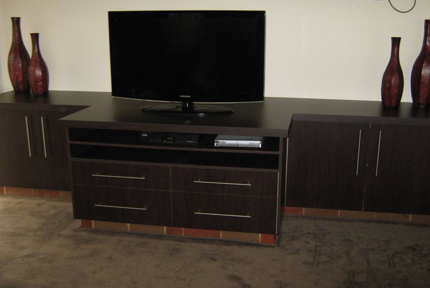 A dark brown entertainment center with a TV, drawers, cabinets, and decorative vases against a cream wall.