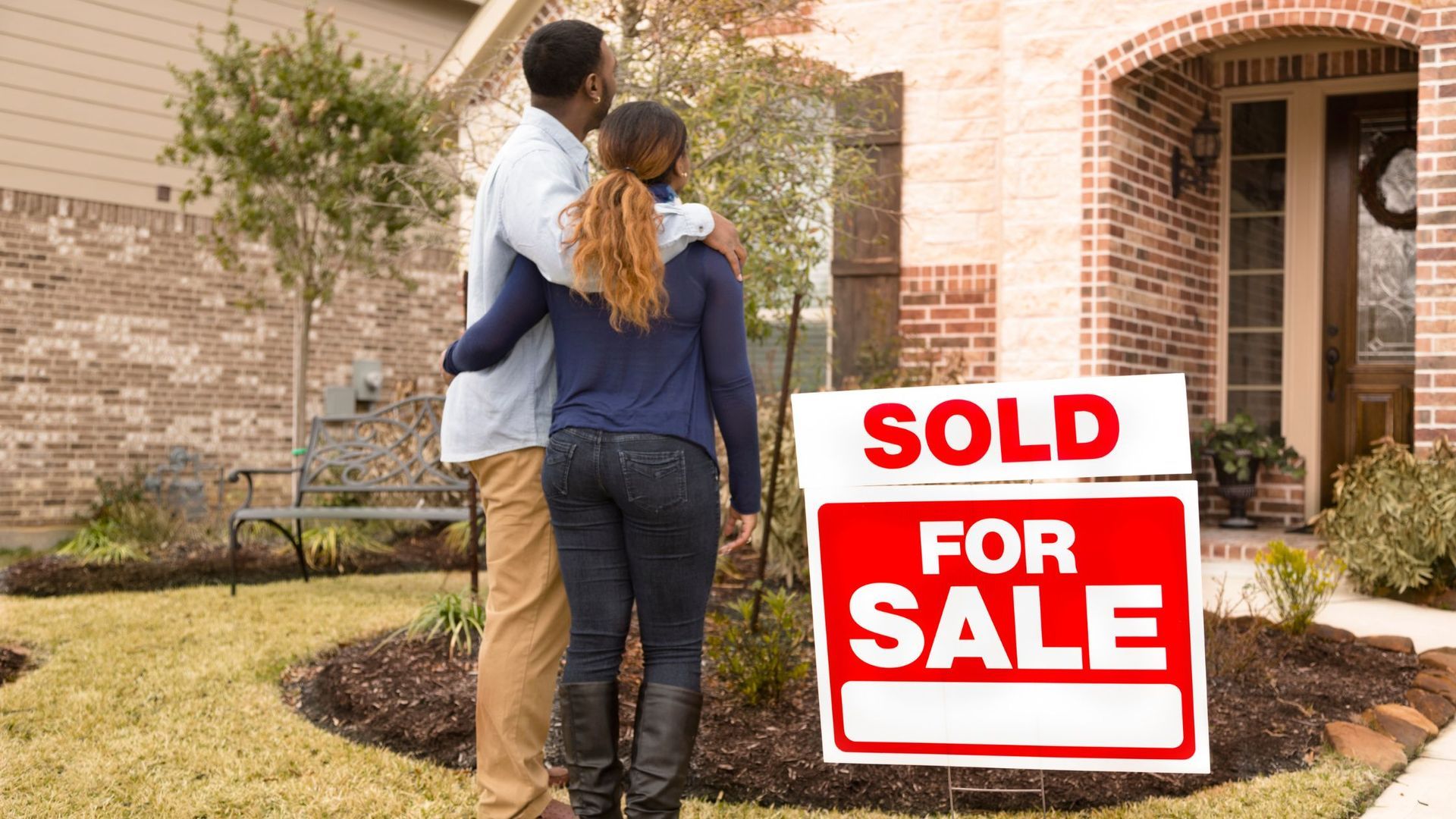 a couple standing in front of their new house with a sold sign on the grass