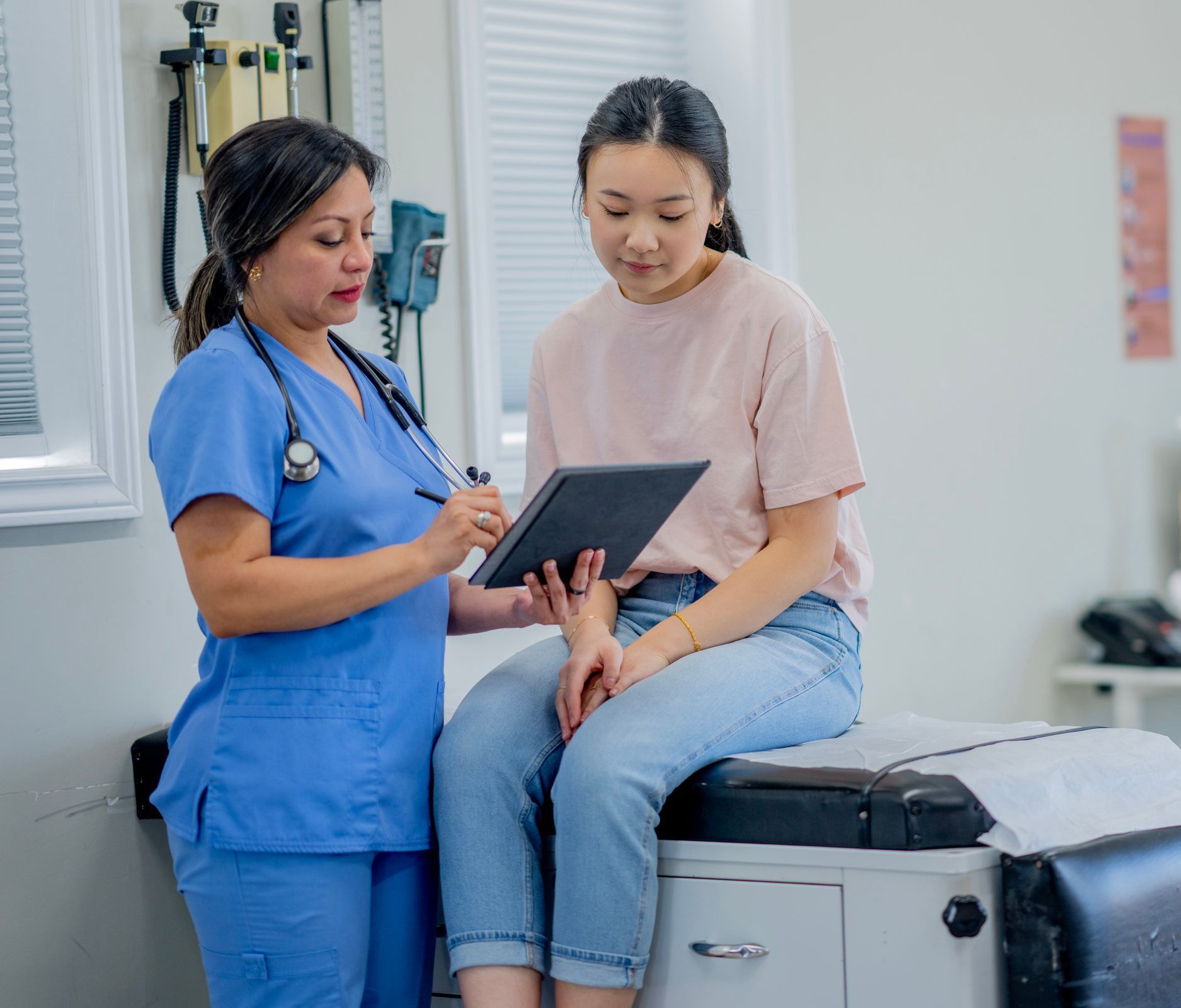 A nurse is talking to a patient who is sitting on a table looking at a tablet.