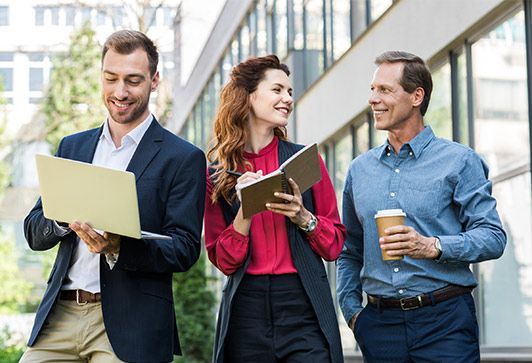 Three coworkers walking outdoors, smiling and talking while holding a laptop, notebook, and coffee cup.