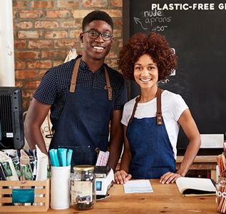 Two smiling cafe staff in aprons behind a wooden counter with reusable cups and supplies