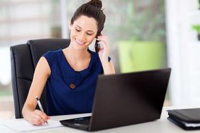 Woman on the phone, smiling at a desk with a laptop and notebook