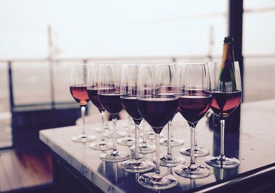 Rows of red wine glasses on a table by a window, with a bottle in the background