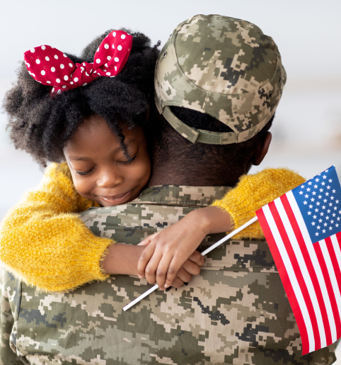 Soldier in camouflage hugs a child with a red bow, holding a small American flag.