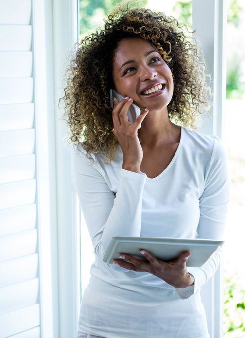 Smiling woman in white top holding a tablet and talking on a phone by a bright window