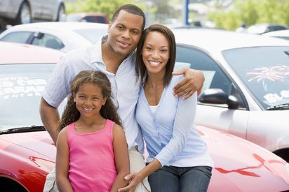 Family of three smiling in front of a red car at a car dealership