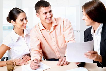 Three coworkers meeting at a table, one holding documents and discussing them in a bright office