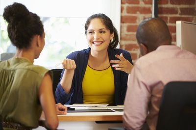 Three people in a meeting, smiling and talking around a table in a bright office.