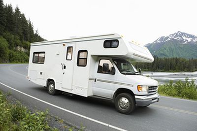 White RV driving on a mountain road beside a river, with pine forest and snowy peaks in the background