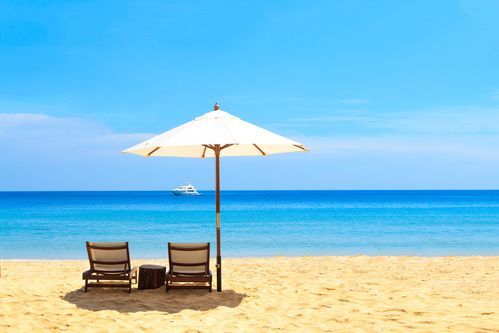 Two lounge chairs under a white beach umbrella on a sunny sandy beach with blue ocean and sky