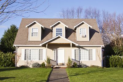 Two-story beige house with brown roof, white trim, and a front lawn under a clear blue sky