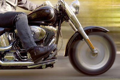 Close-up of a chrome motorcycle riding on a road, with rider’s jeans-clad leg visible.
