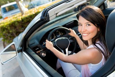 Woman smiling while driving a car, seen from the open window with hands on the wheel.