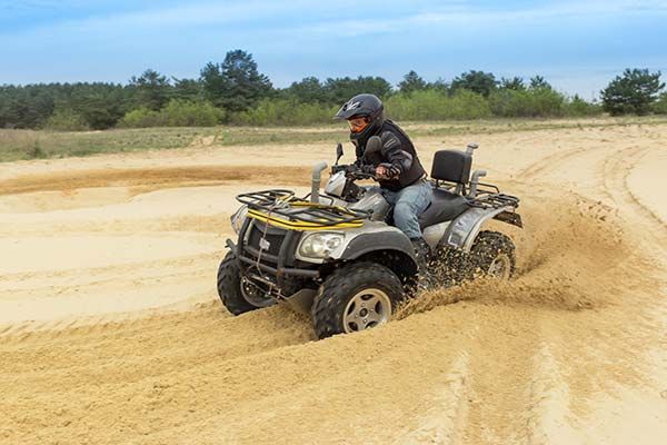 Person riding a yellow ATV through sand, kicking up a cloud of dust in an open off-road area