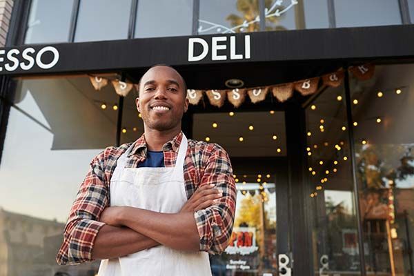 Smiling deli worker in apron standing outside a deli storefront with string lights