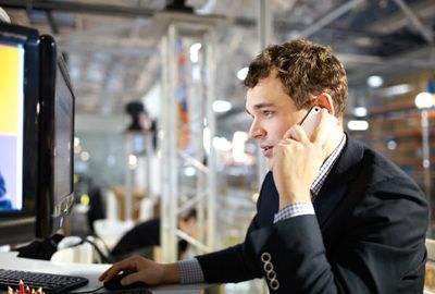 Man in a dark suit on a phone at a desk, working on a computer in an industrial office space