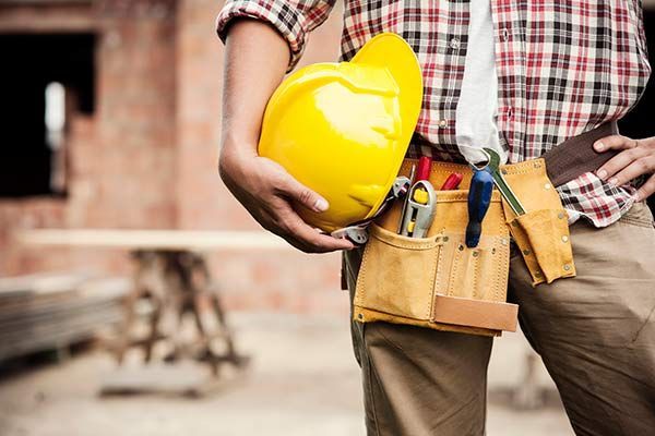 Construction worker holding a yellow hard hat, with tool belt and plaid shirt at a worksite