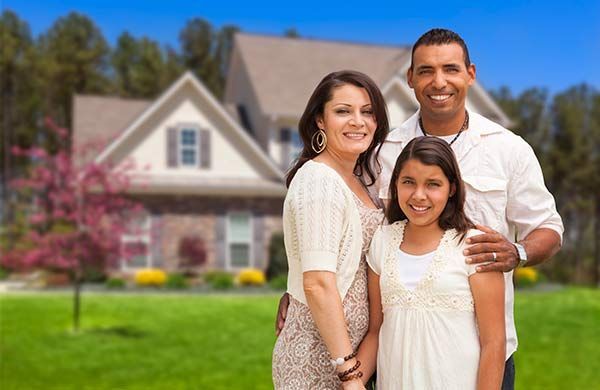 Family smiling in front of a suburban house with a green lawn and pink flowering tree.