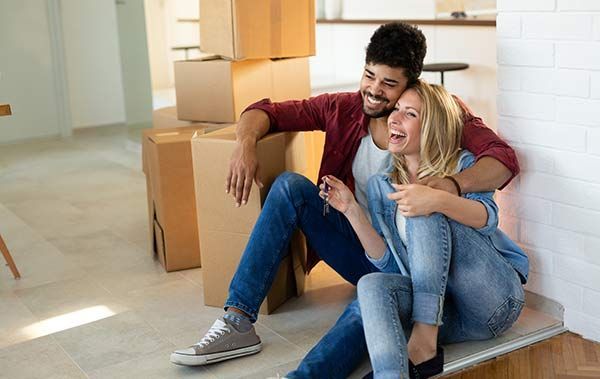 Smiling couple sitting on a floor in a bright room with stacked moving boxes