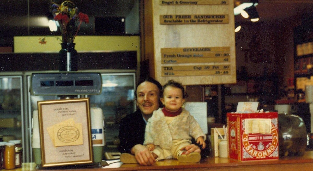 Two people behind a counter with framed signs and posters in a shop or café.