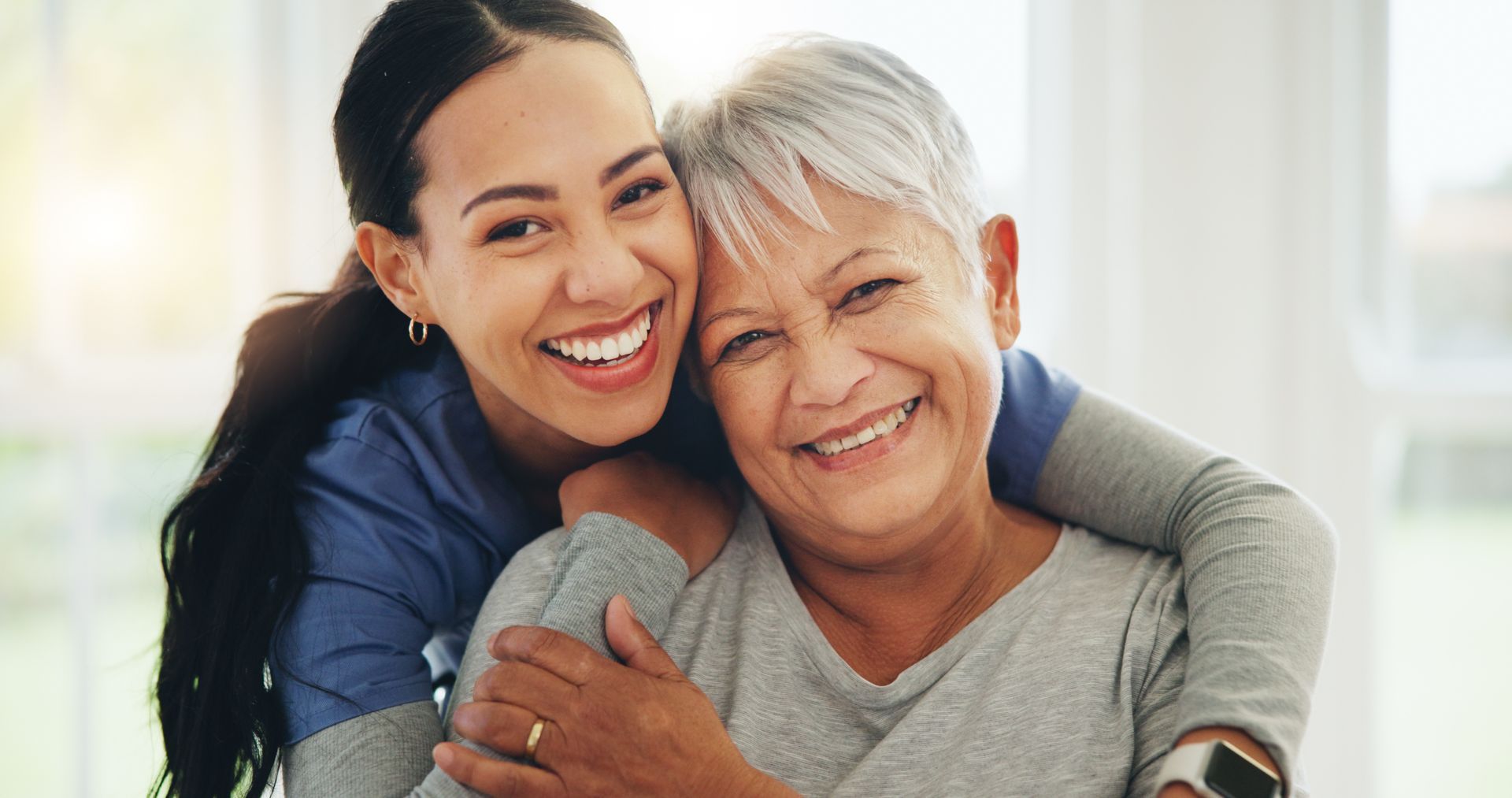 Two smiling people embracing indoors, with a younger person hugging an older person from behind. Two smiling people embracing indoors, with a younger person hugging an older person from behind.