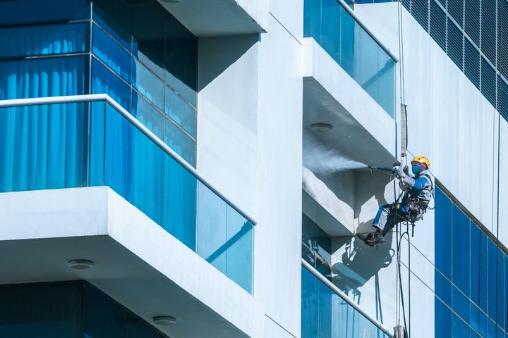 Worker wearing safety harness washes glass facade at height on modern high rise building