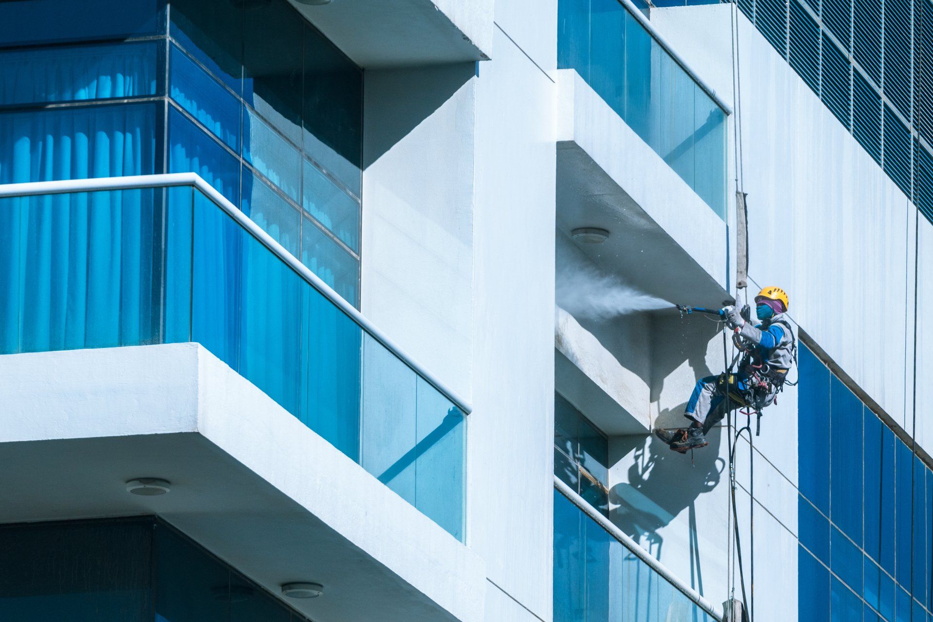 Worker wearing safety harness washes glass facade at height on modern high rise building.