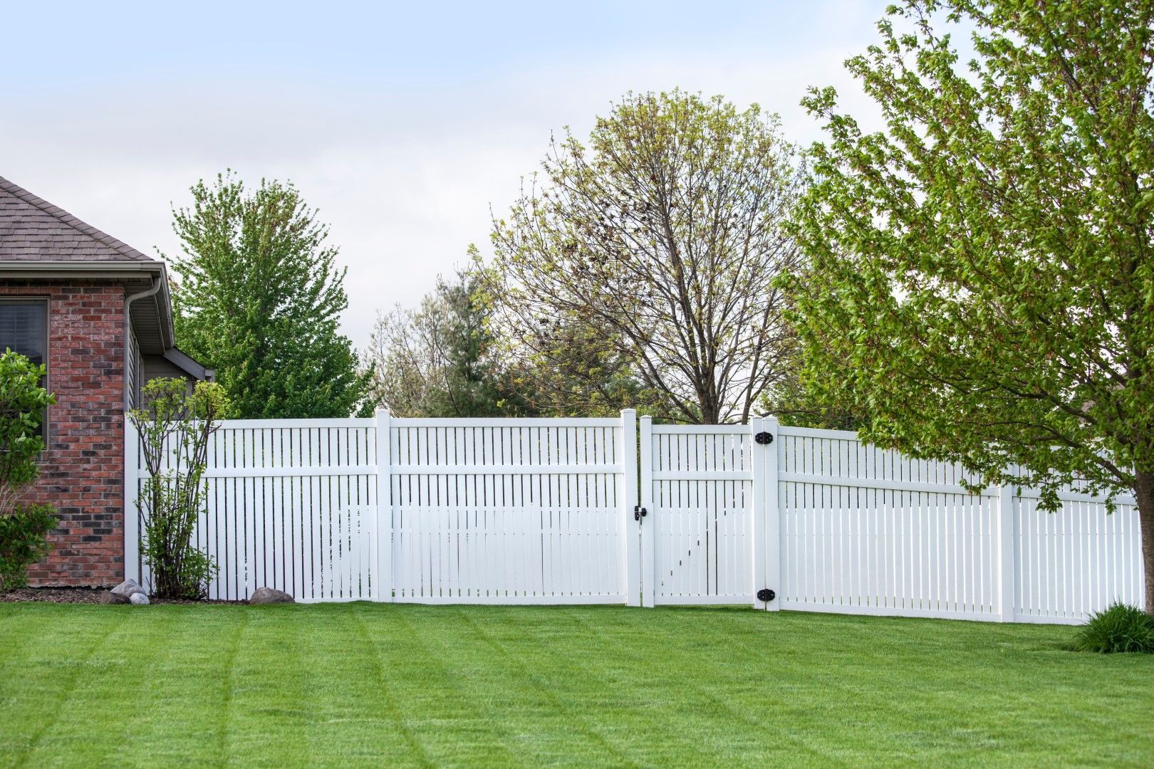 White picket fence around a green lawn, near a brick house and trees.