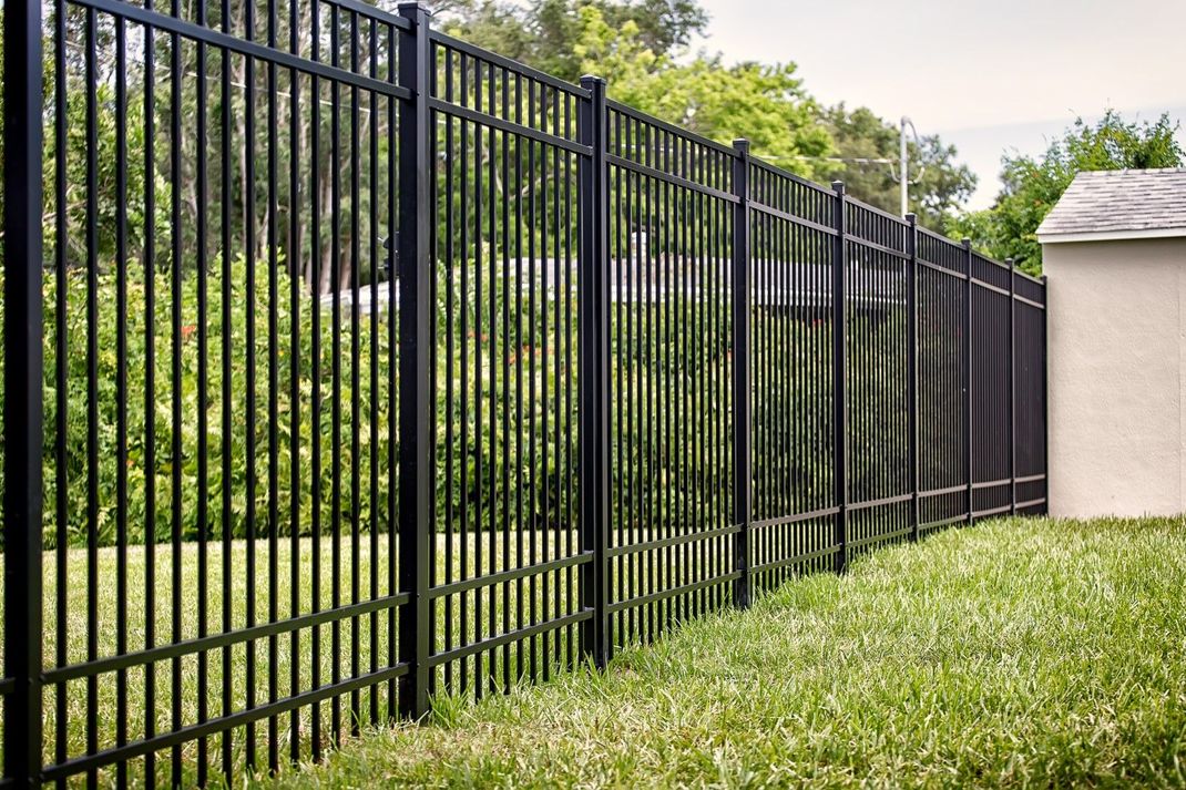 Black metal fence in front of green grass, with trees in the background and part of a building on the right.