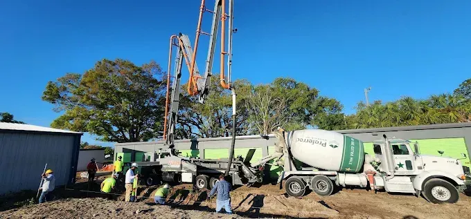 Construction site with concrete pump, mixer truck, workers, and buildings under a blue sky.