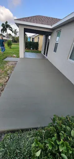 A gray concrete patio next to a house with a green lawn and bushes. Cloudy blue sky.