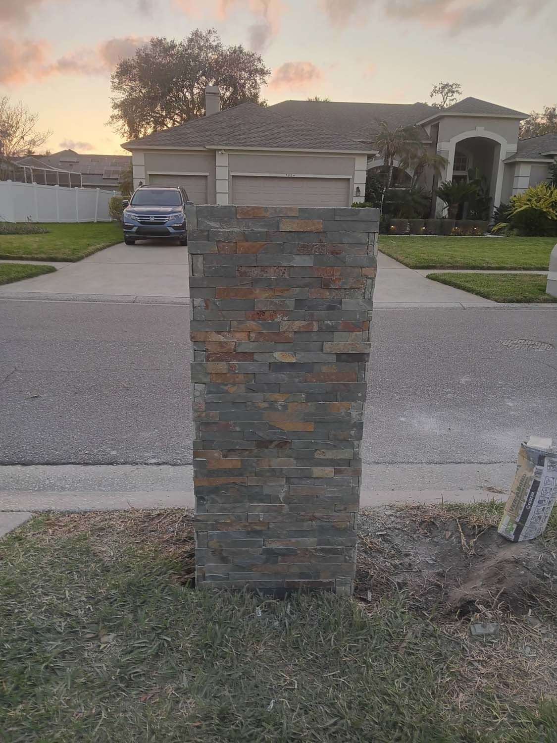 Stone-covered mailbox pillar in front of a house, set in grass beside a road. A car is parked in the driveway.