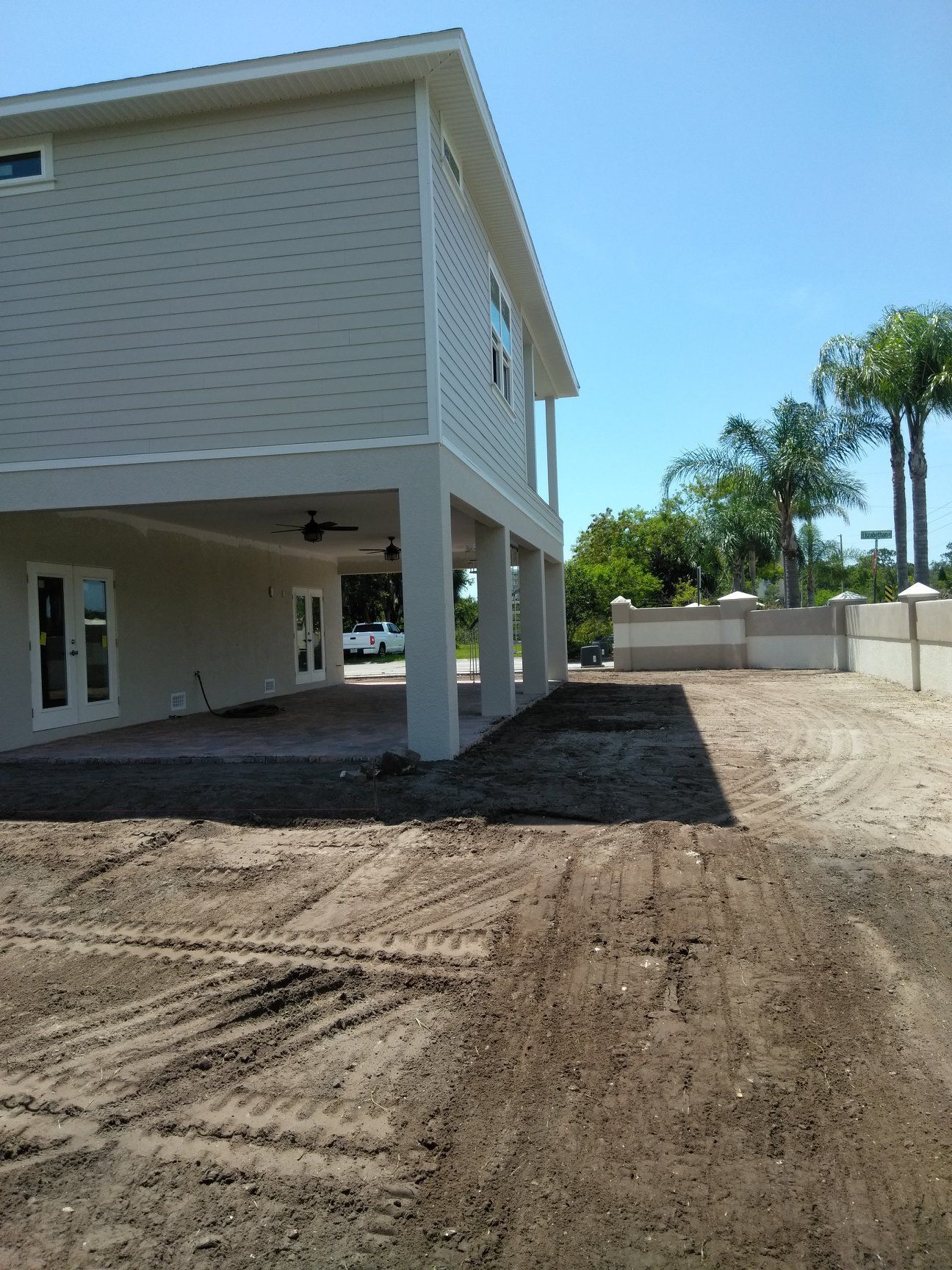 Two-story beige house with a patio under the structure; dirt lot and a wall with palm trees are visible. Two-story beige house with a patio under the structure; dirt lot and a wall with palm trees are visible.