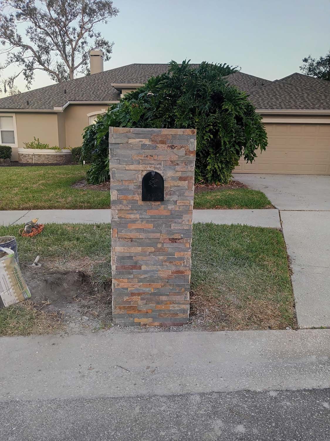 Stone-covered mailbox in front of a tan house with a green lawn and a driveway on a sunny day.