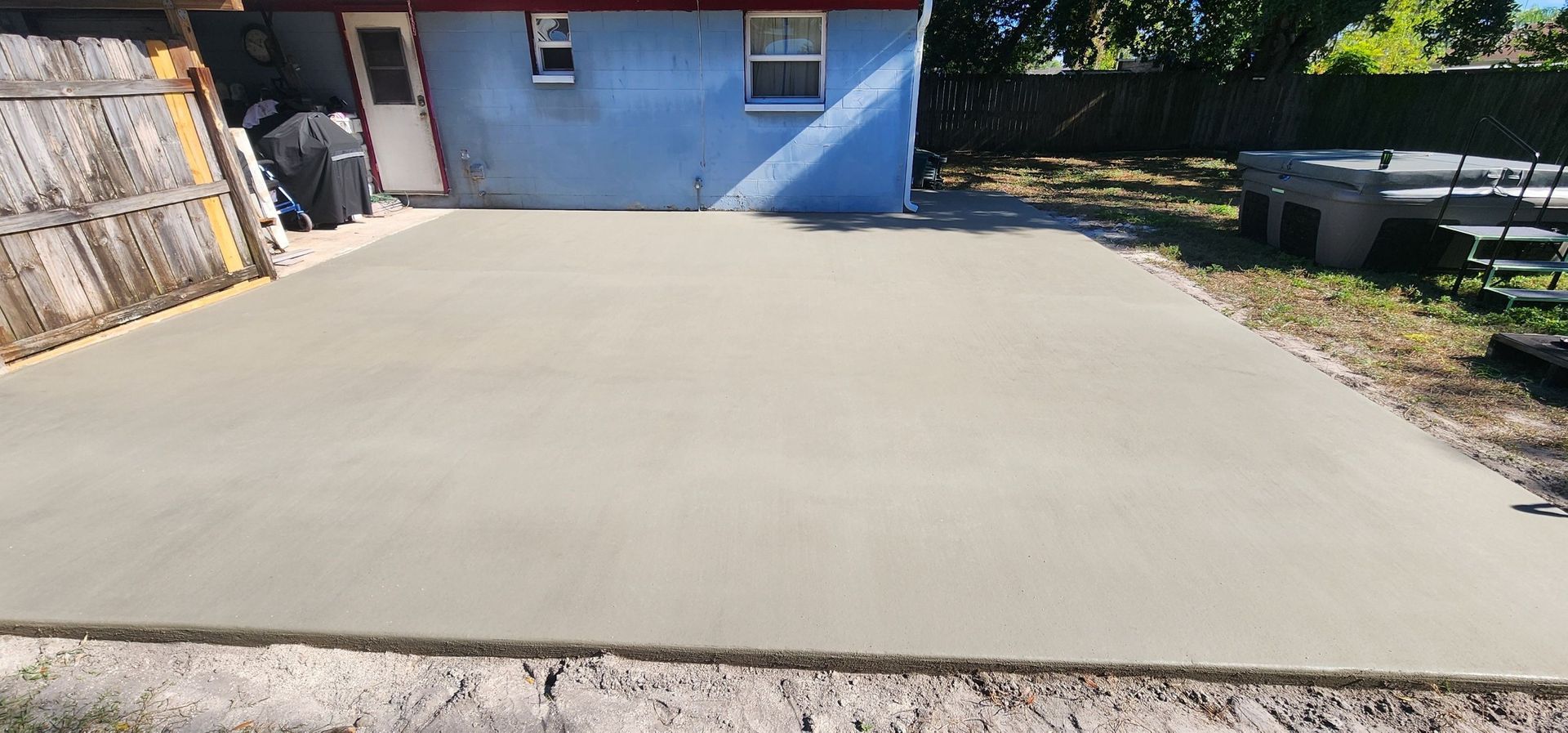 Newly poured concrete patio, adjacent to a blue house and wooden fence, surrounded by grass. Newly poured concrete patio, adjacent to a blue house and wooden fence, surrounded by grass.