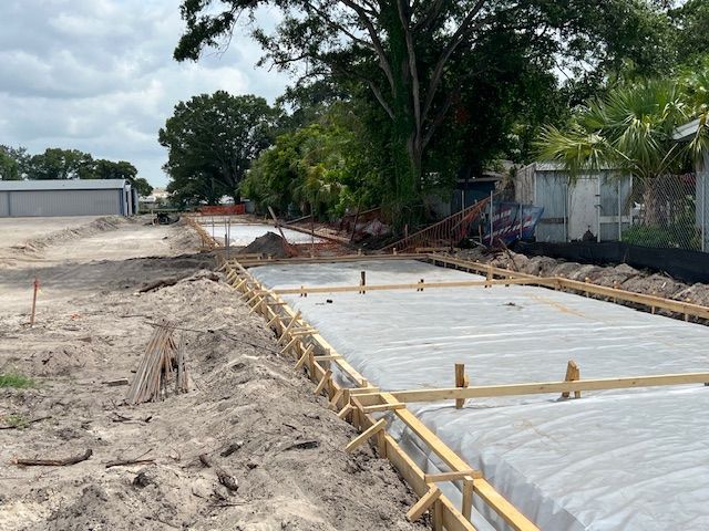 Construction site with wooden forms and gray concrete, next to a sandy area and a building under a cloudy sky. Construction site with wooden forms and gray concrete, next to a sandy area and a building under a cloudy sky.