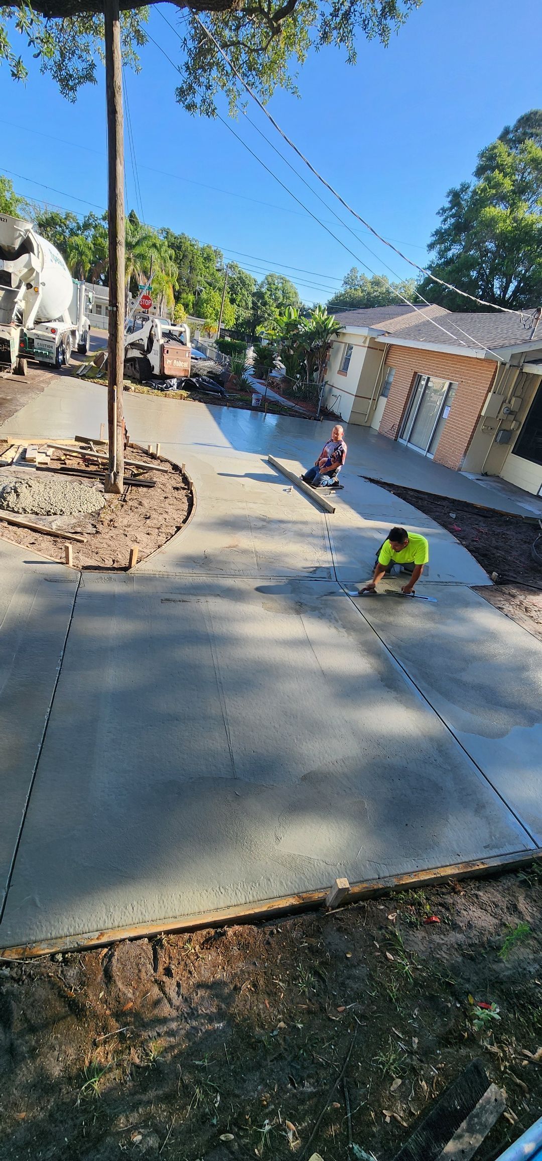 Workers smoothing a fresh concrete driveway. Blue sky, a house, and a truck are in the background. Workers smoothing a fresh concrete driveway. Blue sky, a house, and a truck are in the background.