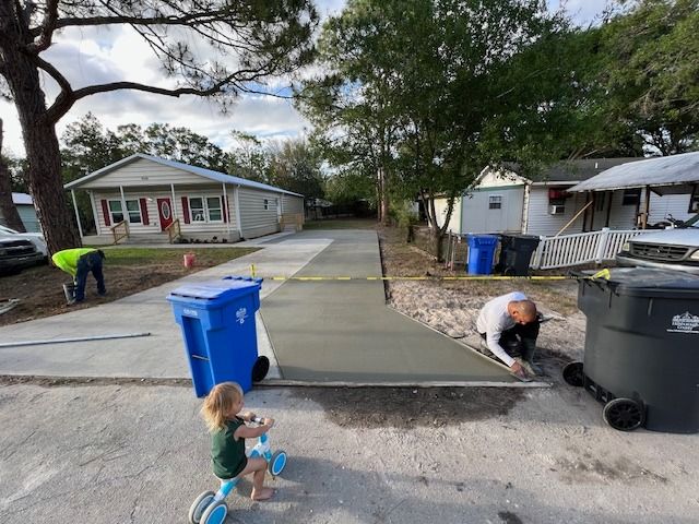 Workers finishing a concrete driveway on a residential street; child on a tricycle nearby. Workers finishing a concrete driveway on a residential street; child on a tricycle nearby.